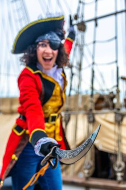 Neşeli bir korsan gibi giyinmiş, göz bandı ve şapka takan, ağzı açık bir kılıç tutan, arka planda donanımlı bir gemi güvertesinde şenlikli bir karnaval olayını kutlayan bir kadın.