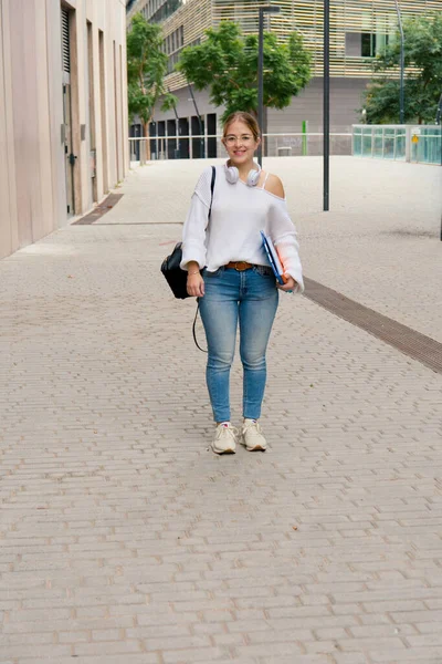 Üniversite kampüsü yolunda yürürken kameraya gülümseyen genç bayan öğrenci, not defterleri, sırt çantaları ve kulaklıklar, günlük kot pantolonlar ve gözlükler, parlak şehir akademik ortamı