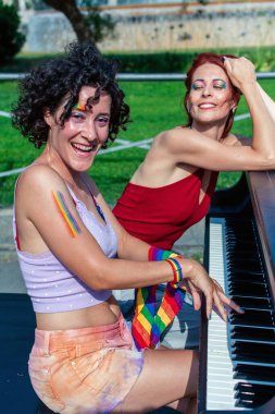 Igbtq gururunu gökkuşağı makyajı ve aksesuarlarıyla ifade eden iki neşeli kadın, biri aktif olarak piyano çalarken diğeri parkta onun yanında mutlu bir şekilde gülümsüyor.