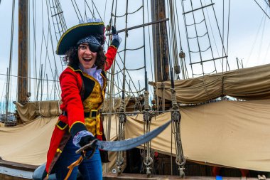 Korsan kostümlü bir kadın, göz bandı ve kılıçla, gülümseyen ve elini kaldıran, tarihi bir ahşap yelkenli gemisinin güvertesinde karnaval kutlaması için duran