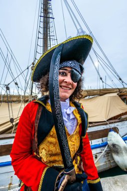 Korsan kostümü giyen, bikorne şapkalı, göz bandı takan ve kılıç kuşanmış bir insan, şenlikli bir karnaval sırasında güvertede neşeli bir ifadeyle poz veriyor.