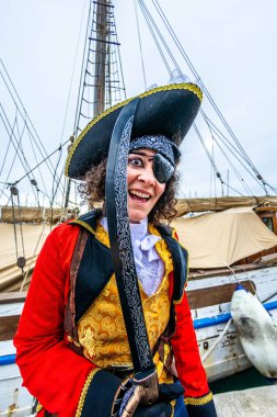 Korsan kostümü giymiş enerjik bir insan, göz bandı takıyor ve elinde palayla, bir yelkenli gemisinin güvertesinde mutlu bir şekilde poz veriyor, karnavalı kutluyor.