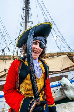 Heyecanlı korsan göz bandı, bandana ve trikorne şapka, kırmızı ceket ve altın yelek, yelkenliye kılıç sallamak, kutlama yapmak ve güvertede neşe içinde poz vermek.
