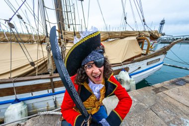 Şapka, göz bandı ve kılıçlı korsan kostümlü bir adam şenlikli bir karnaval sırasında kalabalık bir limana yanaşmış geleneksel bir yelkenli gemisinin önünde neşeyle poz veriyor.