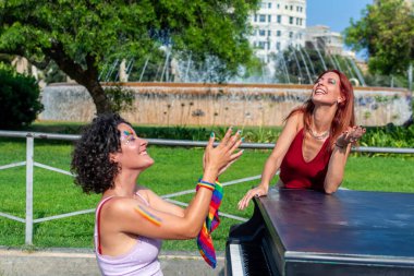 Gülümseyen iki kadın gururu ve kapsamayı gökkuşağı yüz boyası ve bilekliklerle kutluyorlar. Parktaki bir çeşmenin yanındaki açık hava müzik gösterisine katılıyorlar.