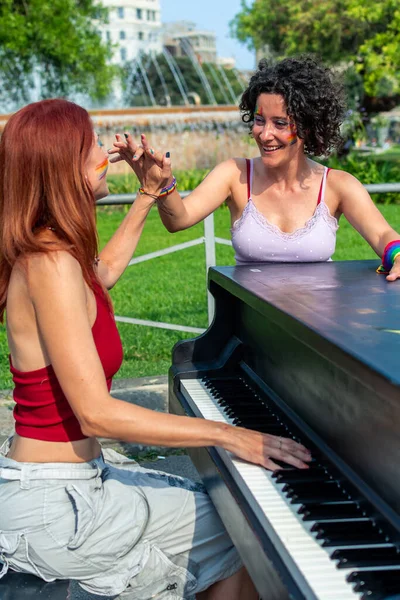 Açık hava piyanosu çalarken neşe ve birlik ifade eden iki neşeli kadının yüzleri gökkuşağı renkleriyle boyanmış, halka açık bir parkta lgbtq gururunu ve çeşitliliğini kutluyorlar.