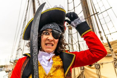 Korsan gibi giyinmiş, şapka takan, göz bandı takan, kırmızı ceket giyen ve elinde kılıç tutan biri, tarihi bir yelkenli gemisinde vahşi bir yüz ifadesi sergiliyor.