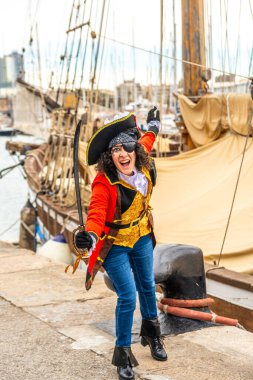 Korsan kaptanı gibi giyinmiş bir kadın, bir kılıç tutuyor ve kutlama jesti olarak kolunu kaldırıyor. Karnaval şenlikleri sırasında arka planda büyük bir yelkenli gemisiyle rıhtımda duruyor.