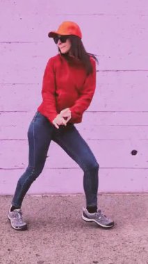 Modern gündelik giysiler içinde genç kaygısız bir kadın canlı pembe beton bir duvara karşı hip hop dansı yaparken eğleniyor, enerjik hareketlerini ve pozitif tutumunu sergiliyor.