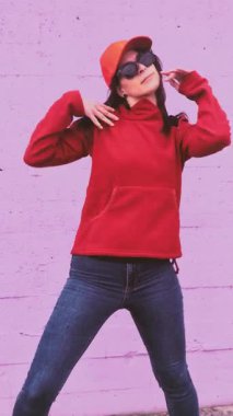 Günlük kıyafet giymiş genç bir kadın renkli bir şehir duvarına karşı hip hop dansı yapıyor, enerjik ve anlamlı hareketler sergiliyor kendine güvenen ve oyuncu bir tavırla.