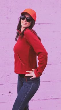 Kırmızı kapüşonlu, şapkalı, hip hop ve sokak tarzı dans eden enerjik genç bir kadın canlı mor bir duvara yaslanıyor, performans sanatı yoluyla kendine güveni ve modern şehir yaşam tarzını ifade ediyor.