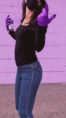 Yüz maskesi, kep ve mor eldivenler takan genç bir kadın, mor bir duvara karşı sprey boya kutusu kullanırken enerjik bir şekilde dans ediyor, kendini sokak sanatı ve hareketleriyle ifade ediyor.