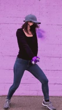 Yüz maskesi, kep ve mor eldivenler takan genç bir kadın dans ediyor ve mor bir duvara karşı sprey boyayla graffiti yapıyor, modern sokak sanatında şehir isyanı ve yaratıcılığını ifade ediyor.