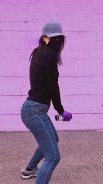 Maskeli ve eldivenli genç kadın grafiti sanatçısı elinde sprey kutusuyla mor bir duvara karşı dans ediyor, isyankar ve yaratıcı sokak sanatını şehir ortamında sergiliyor.