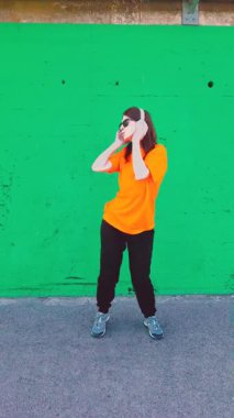 Güneş gözlüklü ve kulaklıklı genç enerjik kadın müzik dinliyor ve beton bir duvara karşı dans ediyor, ritmin tadını çıkarıyor ve mutlu ve kaygısız hissediyor.