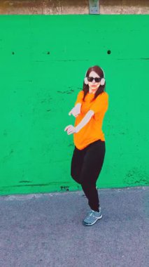 Kulaklık takan ve güneş gözlüğü takan mutlu genç bir kadın güçlü yeşil bir duvarın önünde hip hop dansı yaparak modern şehir dansı hareketleriyle özgürlüğünü ve neşesini ifade ediyor.