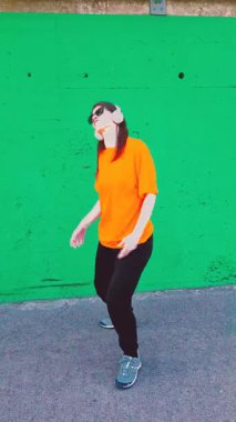 Enerjik bir kadın kulaklık takıyor ve güneş gözlüğü takıyor canlı yeşil beton bir duvara karşı özgürce dans ediyor, müziğin tadını çıkarıyor ve modern dans hareketleriyle mutluluğunu ifade ediyor.