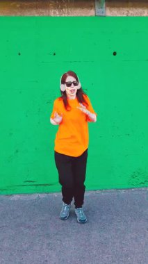 Kulaklık ve güneş gözlüğü takan zarif genç bir kadın canlı yeşil beton bir duvara karşı neşeyle dans ediyor. Modern dans hareketleriyle özgürlüğü ve mutluluğu ifade ediyor.