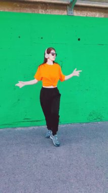 Enerjik bir kadın, kulaklık takıyor ve güneş gözlüğü takıyor canlı yeşil bir şehir duvarına karşı sokak dansı yapıyor, müzikten hoşlanıyor ve modern koreografiyle kendini ifade ediyor.