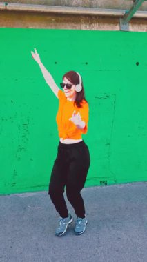 Neşeli bir kadın, kulaklık takıyor ve güneş gözlüğü takıyor enerjik bir şekilde dans ediyor ve parlak yeşil bir şehir duvarına karşı eğleniyor, müzikten hoşlanıyor ve dışarıda hareketlerle neşeli ruh halini ifade ediyor.