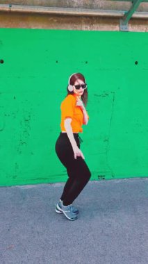 Canlı yeşil bir duvara karşı neşeli bir şekilde dans eden, kulaklık takan ve güneş gözlüğü takan neşeli genç bir kadın, ritme göre hareket ediyor ve saf mutluluk ve özgürlüğü ifade ediyor.