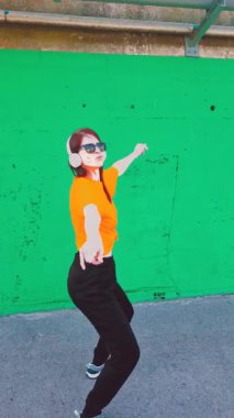 Güneş gözlüklü ve kulaklıklı enerjik mutlu kadın müzik dinliyor, canlı yeşil bir duvarın karşısında özgürce dans ediyor ve coşkulu, spontane hareketlerle saf neşe ve heyecan ifade ediyor.