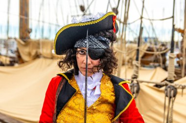 Siyah kıvırcık saçlı, tarihi kostümlü, yüzünde küçük bir kılıçla bir gemi güvertesinde göz bandı ve tricorn şapka takan bir korsan kadın.