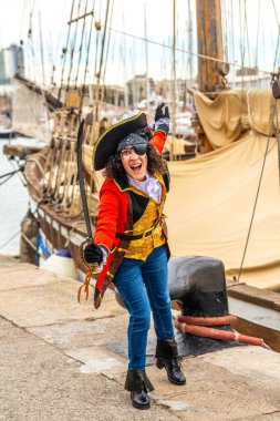 Korsan kostümü giymiş neşeli bir kadın, göz bandı takıyor, şapka takıyor ve kılıç taşıyor, arka planda eski bir yelkenli gemisiyle rıhtımda duruyor, festival sırasında macerayı somutlaştırıyor.