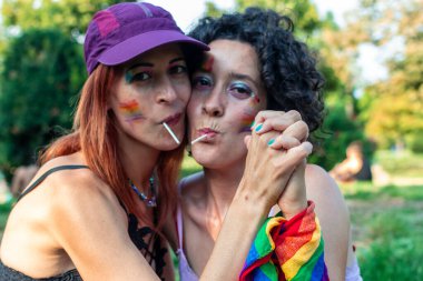 Gökkuşağı makyajlı ve gökkuşağı bayraklı iki kadın, el ele tutuşuyorlar ve lolipoplarını paylaşıyorlar, aşkı, kabulü ve lgbtq 'ı temsil ediyorlar, ve açık havada canlı bir şekilde gururlanıyorlar.