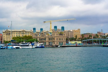 Barselona limanı, liman boyunca demirlemiş tekneleri ve tarihi mimariyi gösteriyor. Bulutlu bir gökyüzüne karşı yeni gelişmelere nezaret eden bir inşaat vinci var.