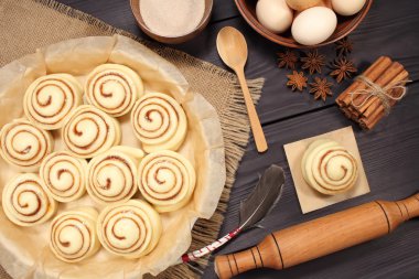 Raw cinnamon rolls prepared for roasting next to eggs and kitchen tools on a background of rustic table of black boards clipart
