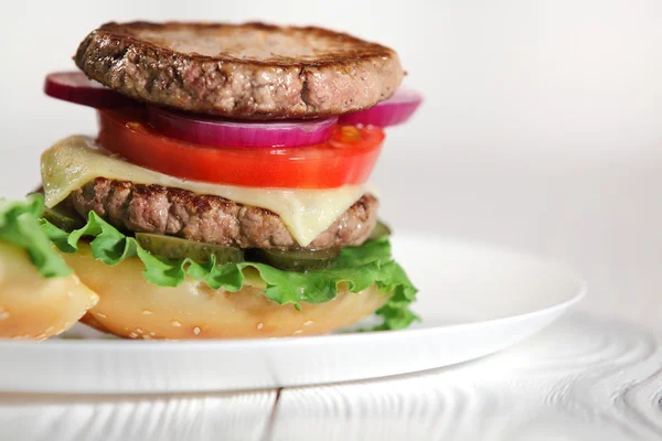 Μεγάλο ζουμερό παραδοσιακά American burger με ψητό κρέας και φέτες φρέσκα λαχανικά κινηματογραφήσεων σε ένα λευκό πιάτο σε ένα ξύλινο τραπέζι. Τρόφιμα και ποτά. Επιλεκτική εστίαση με ρηχό βάθος πεδίου — Φωτογραφία Αρχείου