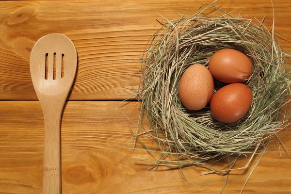 La composición de tres huevos de pollo en un nido de hierba y cuchara de madera acostado en un panel de tableros marrones vintage con espacio libre para la publicidad de texto de la comida o el diseño de menú de restaurante . — Foto de Stock