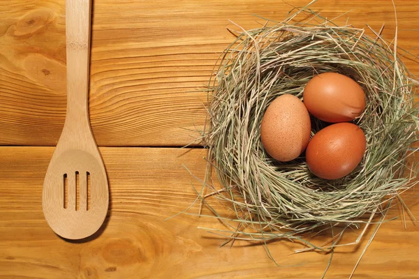 La composición de tres huevos de pollo en un nido de hierba y cuchara de madera acostado en un panel de tableros marrones vintage con espacio libre para la publicidad de texto de la comida o el diseño de menú de restaurante . — Foto de Stock
