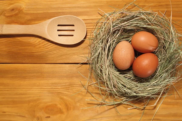 La composizione di tre uova di pollo in un nido da erba e cucchiaio di legno sdraiato su un pannello di tavole marroni vintage con spazio libero per la pubblicità testuale del cibo o del design del menu del ristorante . — Foto Stock