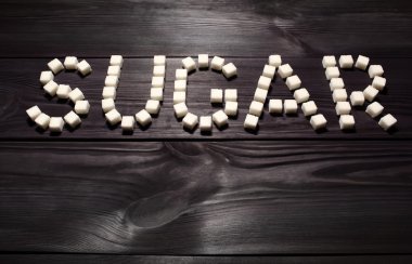 'Şeker' kelimesi, beyaz küp şeker parçalarından oluşmuştur. Arka planda, eski tahta panoların siyah renginde, yüksek kontrastlı 'Sert Işıklandırma'.