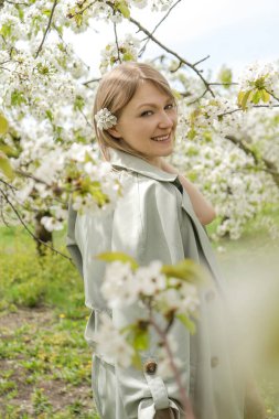 Genç, güzel, neşeli bir kadın güneşli bir bahar gününde güzel bir kadın kıyafetiyle çiçek açan kiraz ağaçlarının altında yürüyor..