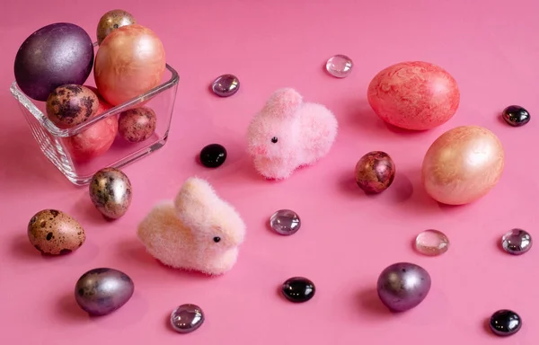 Paskalya. Çok renkli, sedef yumurtalar pembe arka planda ve cam taşlarla süslenmiş küçük bir vazoda bulunur. Yakınlarda iki tavşan var, pembe ve sarı..