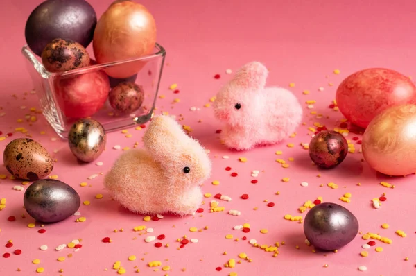 Paskalya. Çok renkli, sedef yumurtalar pembe arka planda ve küçük cam bir vazoda bulunur. Yakınlarda iki tavşan var, pembe ve beyaz..