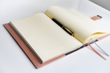 Üzerinde siyah ve altın bir kalem bulunan bej ve sarı çarşaflı ve soluk pembe kapaklı açık bir defter beyaz bir masada duruyor..