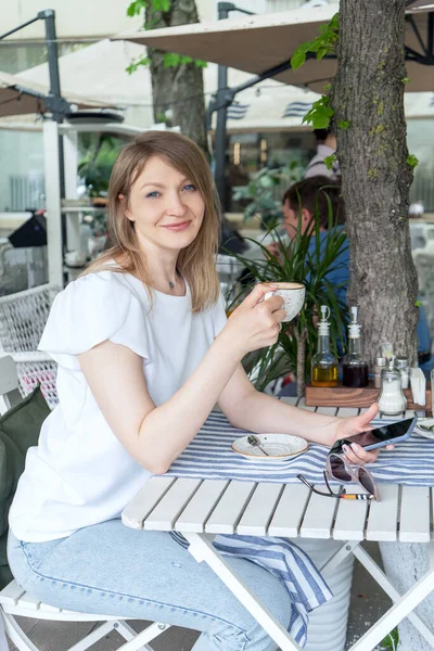 Genç, güzel, neşeli bir kadın bir kafe terasında oturuyor, kahve içiyor ve elinde bir telefon tutuyor..