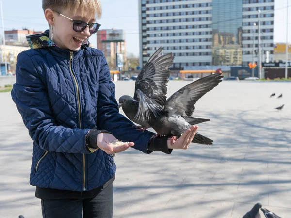 Genç bir kız meydanda duruyor ve güvercinleri ellerinden besliyor, hem de gülerek..