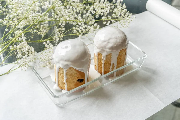 Paskalya. Beyaz kağıt üzerinde, küçük beyaz çiçeklerin yanında, içinde iki Paskalya pastası olan bir vazo var..