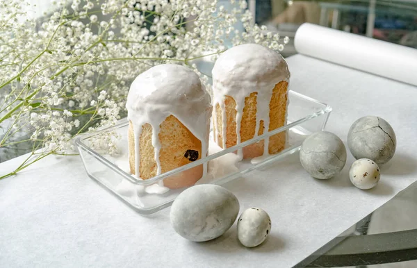 Paskalya. Beyaz kağıt üzerinde, küçük beyaz çiçeklerin yanında, içinde iki Paskalya pastası olan bir vazo var..