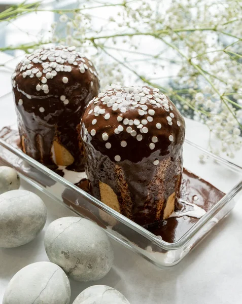 Paskalya. Beyaz kağıt üzerinde, küçük beyaz çiçeklerin yanında, içinde iki Paskalya pastası olan bir vazo var..