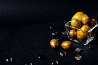 Paskalya tatili. Çok güzel, çeşitli boyutlarda altın yumurtalar siyah bir zemin üzerinde, cam taşlarla süslenmiş küçük cam bir vazoda yatıyor..