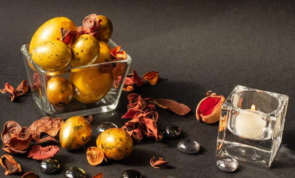 Paskalya tatili. Çeşitli boyutlarda güzel, altın yumurtalar, kurumuş çiçekler ve cam taşlarla süslenmiş, siyah bir zemin üzerinde küçük cam bir vazoda yatıyor. Yakınlarda beyaz mumlu küçük camdan bir şamdan var..