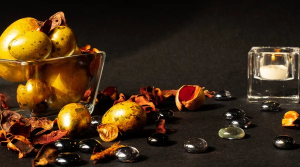 Paskalya tatili. Çeşitli boyutlarda güzel, altın yumurtalar, kurumuş çiçekler ve cam taşlarla süslenmiş, siyah bir zemin üzerinde küçük cam bir vazoda yatıyor. Yakınlarda beyaz mumlu küçük camdan bir şamdan var..