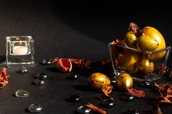 Paskalya tatili. Çeşitli boyutlarda güzel, altın yumurtalar, kurumuş çiçekler ve cam taşlarla süslenmiş, siyah bir zemin üzerinde küçük cam bir vazoda yatıyor. Yakınlarda beyaz mumlu küçük camdan bir şamdan var..