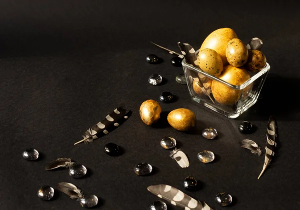 Paskalya tatili. Farklı boyutlarda güzel, altın yumurtalar siyah bir zemin üzerinde, tüylerle ve cam taşlarla süslenmiş küçük cam bir vazoda yatıyor..